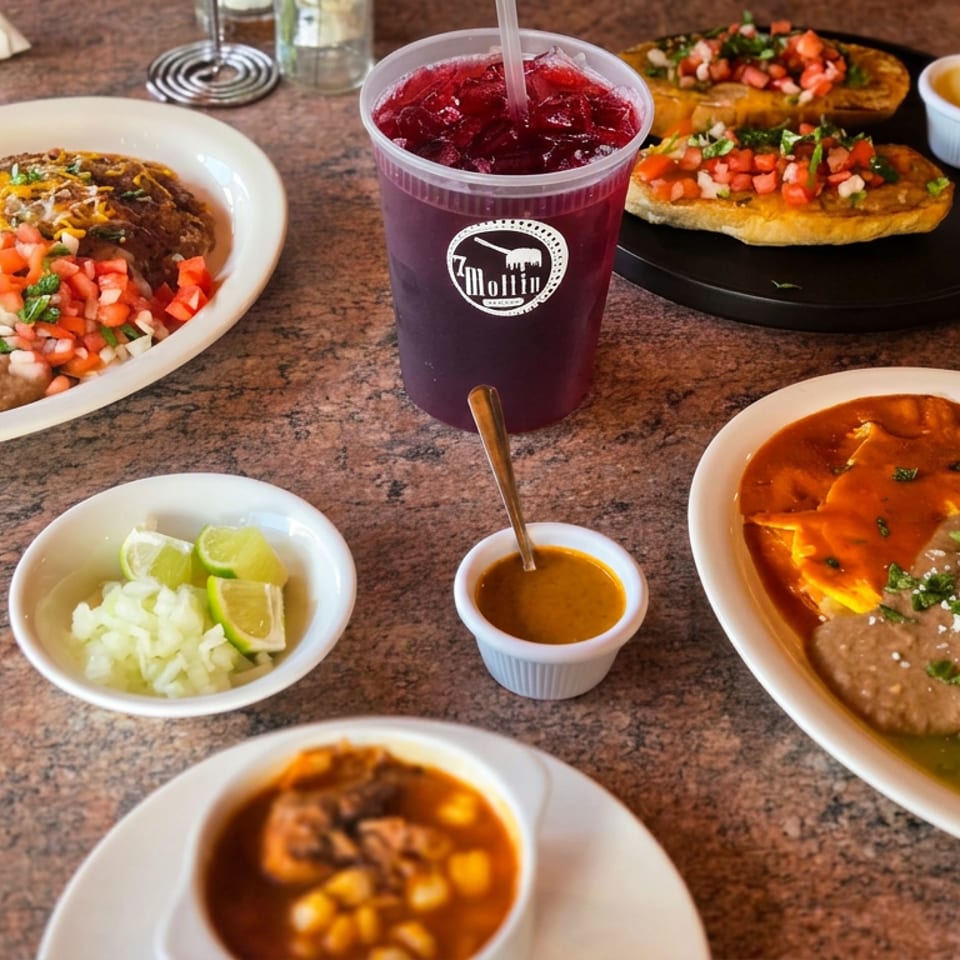Mexican dishes including enchiladas, refried beans, tostadas, salsa, lime, and a purple fruit drink arranged on a restaurant table.