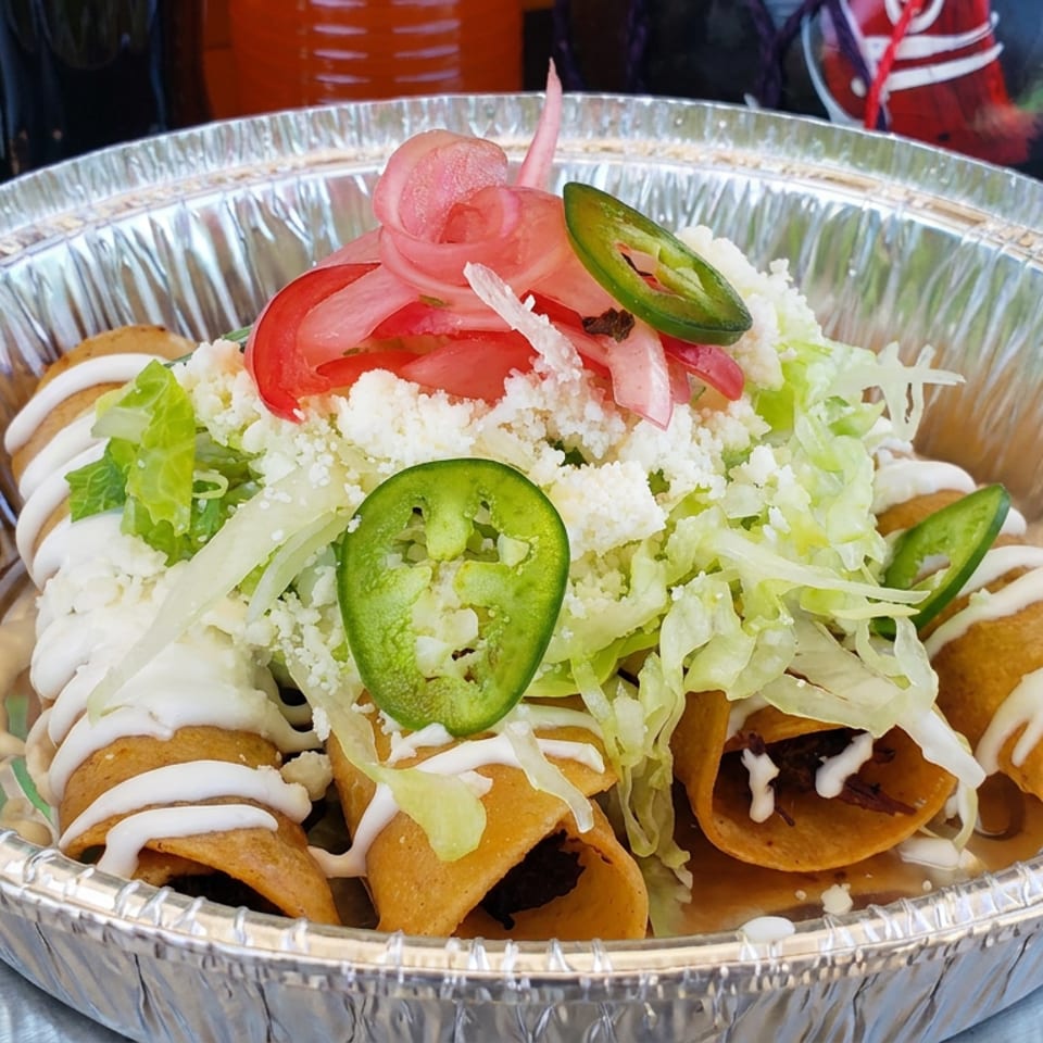 Crispy rolled tacos topped with lettuce, cotija cheese, tomatoes, pickled onions, jalapeños, and drizzled cream in a foil bowl.