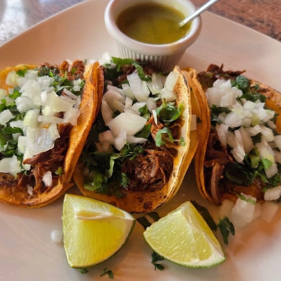 Three tacos topped with chopped onions and cilantro, served with lime wedges and green salsa on a white plate.