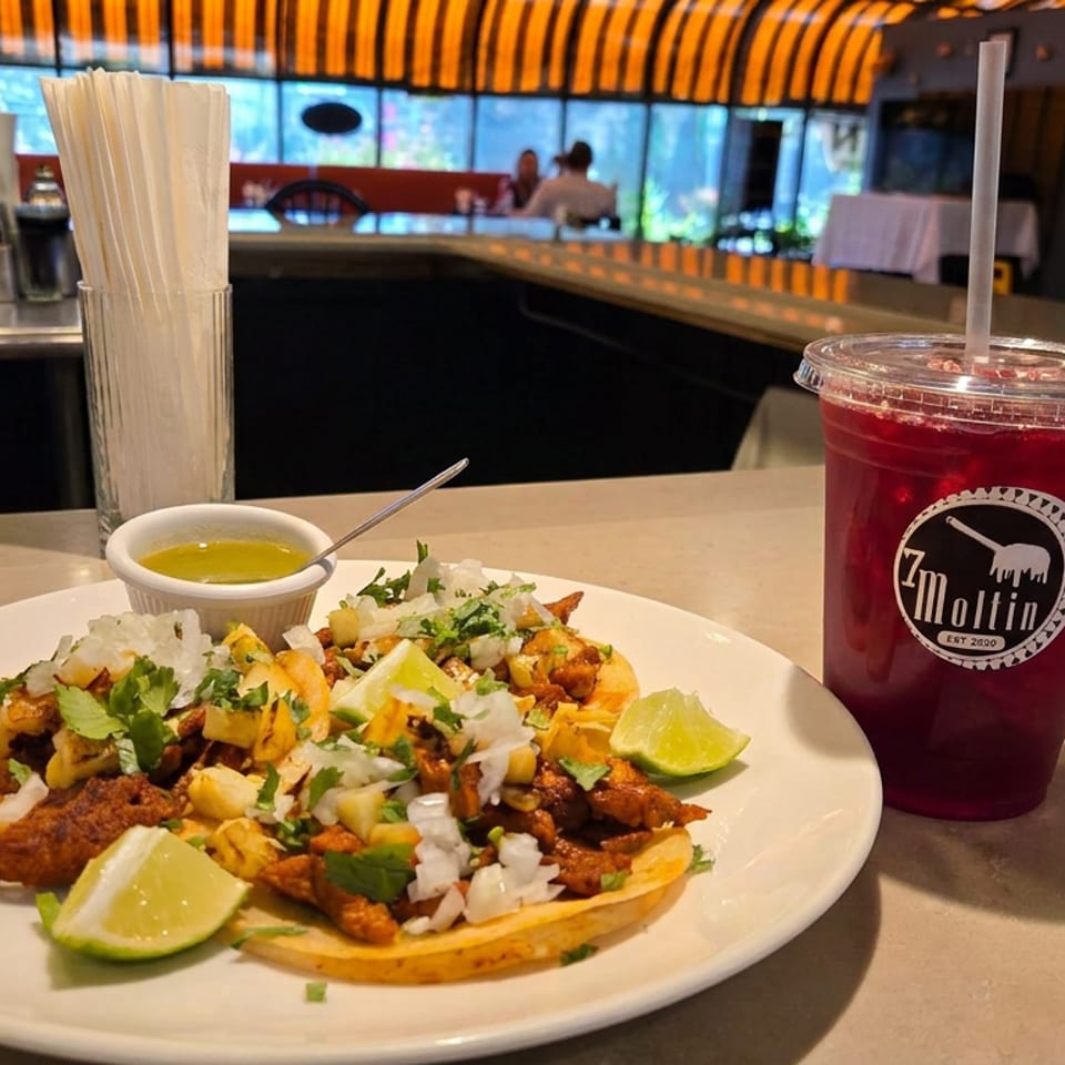 Loaded tacos topped with grilled meat, onions, cilantro, lime wedges, and dipping sauce beside a red drink at a colorful restaurant bar.